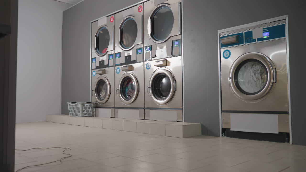 Zoom in on stainless steel washers washing clothes in row, unit by corner, laundry baskets near shelf, digital timers glowing, front load doors spinning, interior service space during wash cycle