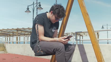 young man sitting on swing in playground on phone