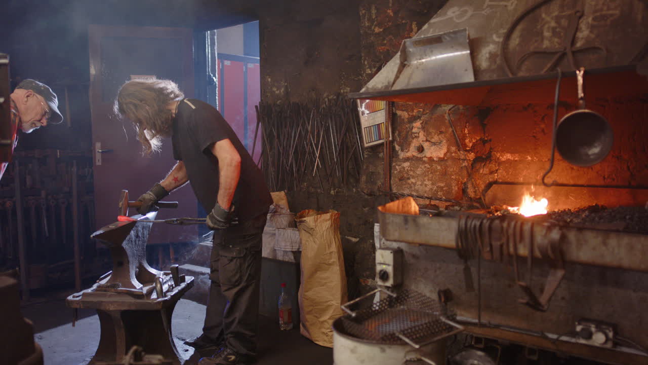 A blacksmith apprentice hammers and is advised by his teacher in the smithy