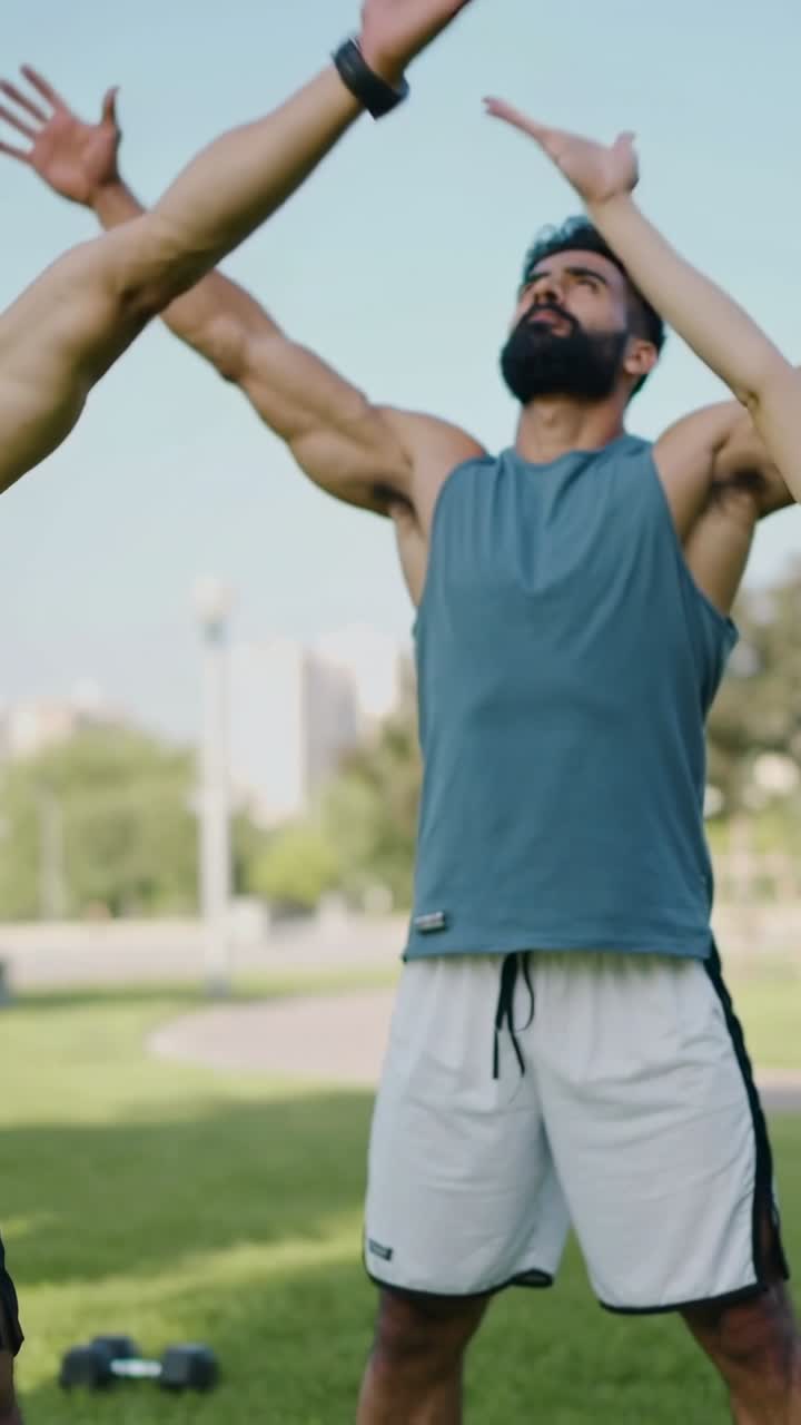 Vertical video: Leading warmup on cue, bearded man in teal tank raising arms in park with dumbbells