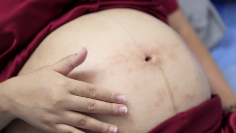 pregnant woman wearing a pair of red silk pajamas is lying down on the bed as she is gently stroking her swollen belly with both of her hands