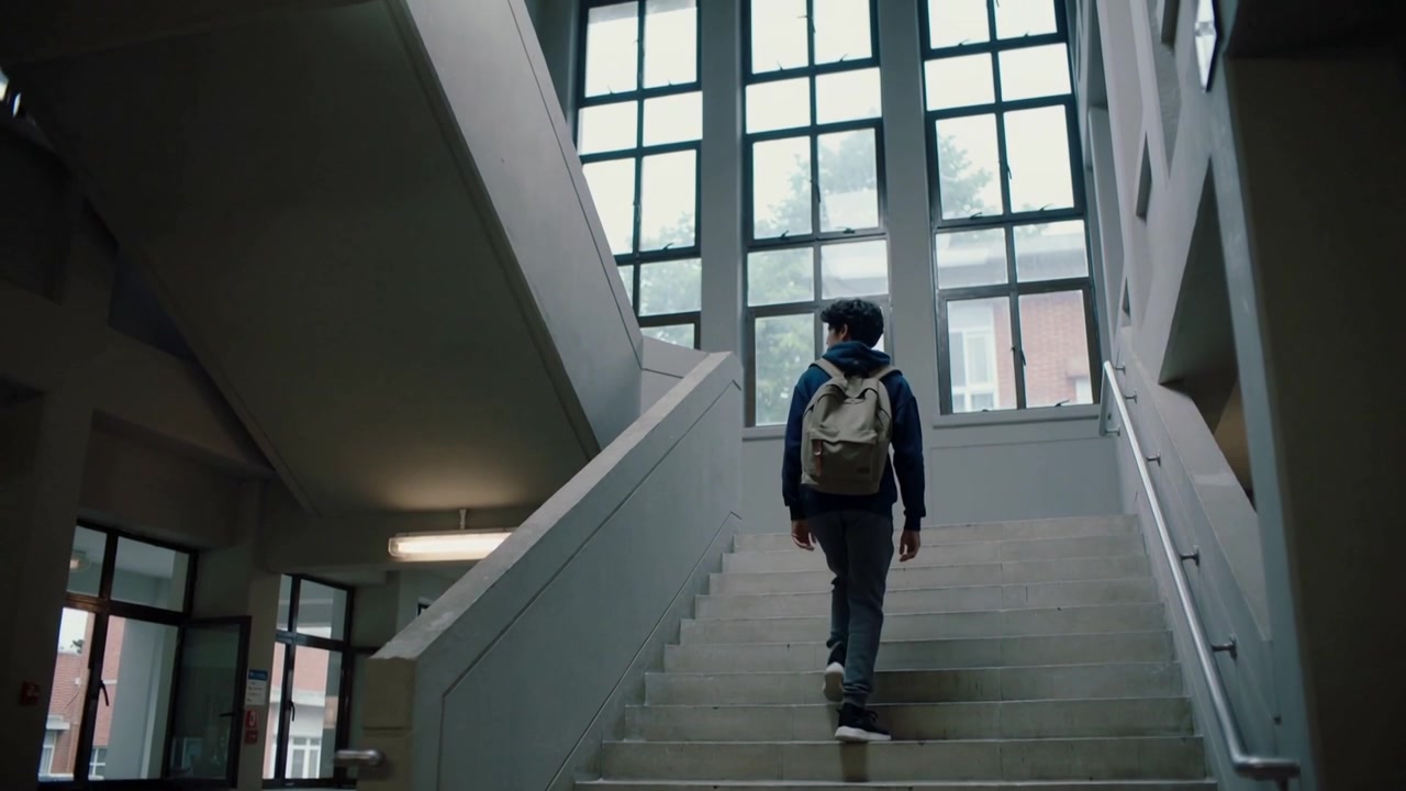 Forlader nederste repos, hætteklædt studerende går op ad trapperne på skolen med en beige rygsæk på vej til undervisning.