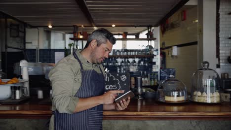 good looking male cafe owner wearing apron, concentrating while scrolling on tablet, standing in funky coffee shop