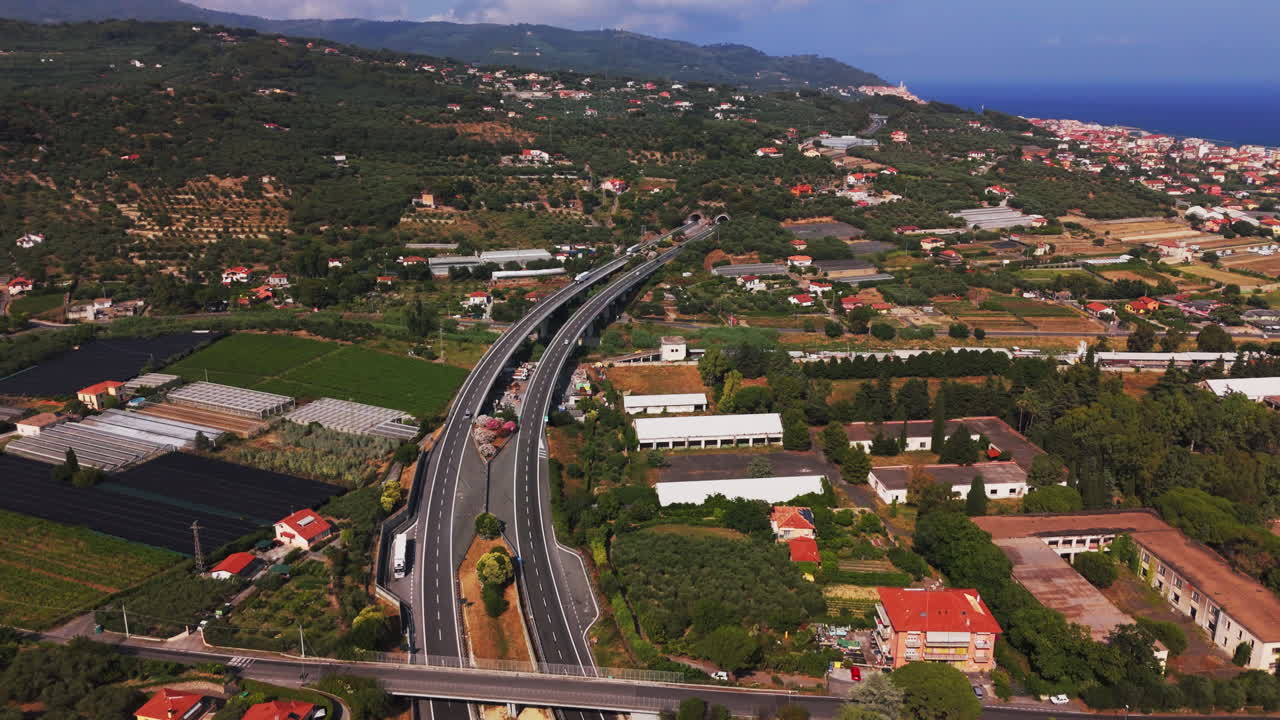 High-altitude drone shot with a panning motion over a coastal area showing roads, tunnel, farmland and residential buildings in Varigotti, Liguria