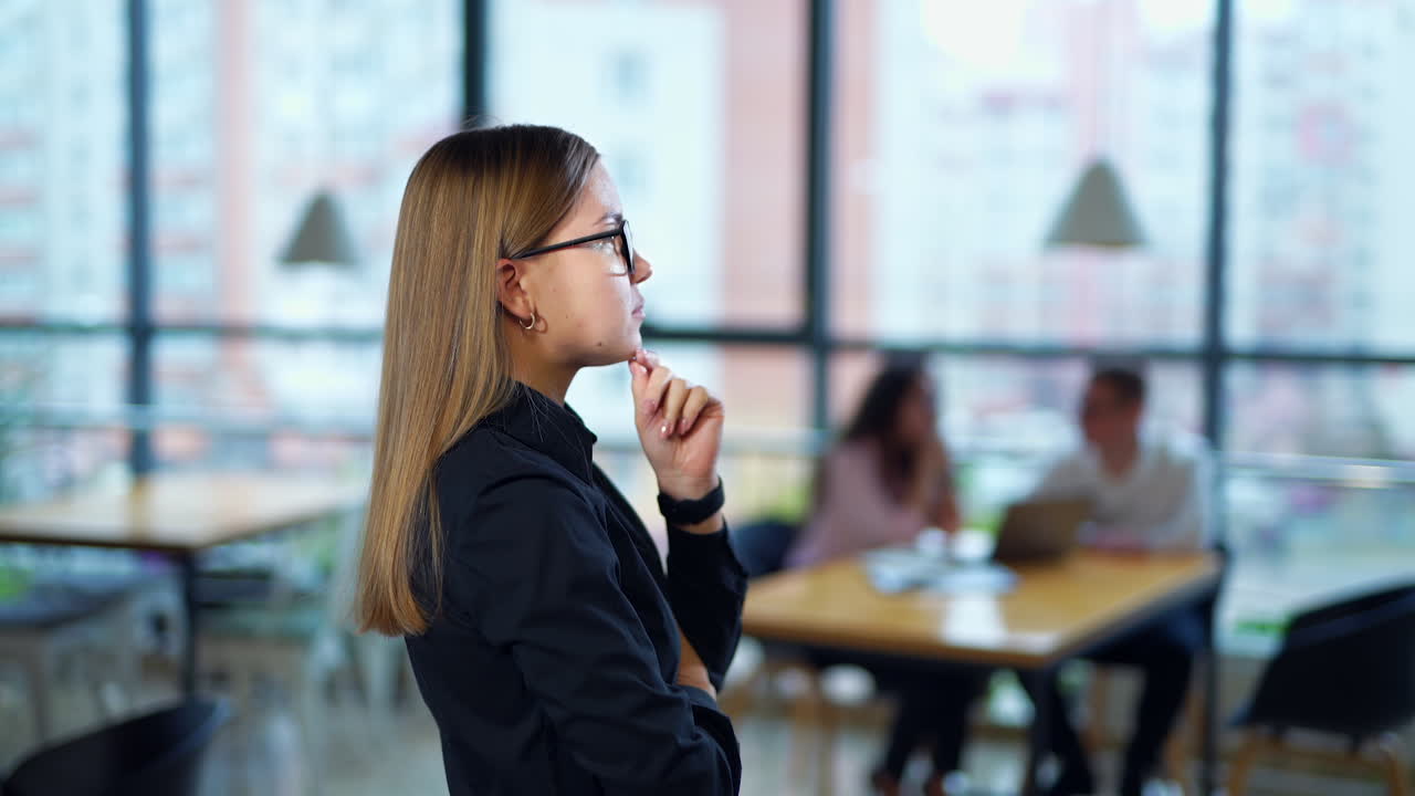 Young female employee standing in office thinking over something. Side view. Two people talking at the table in blur at the background.