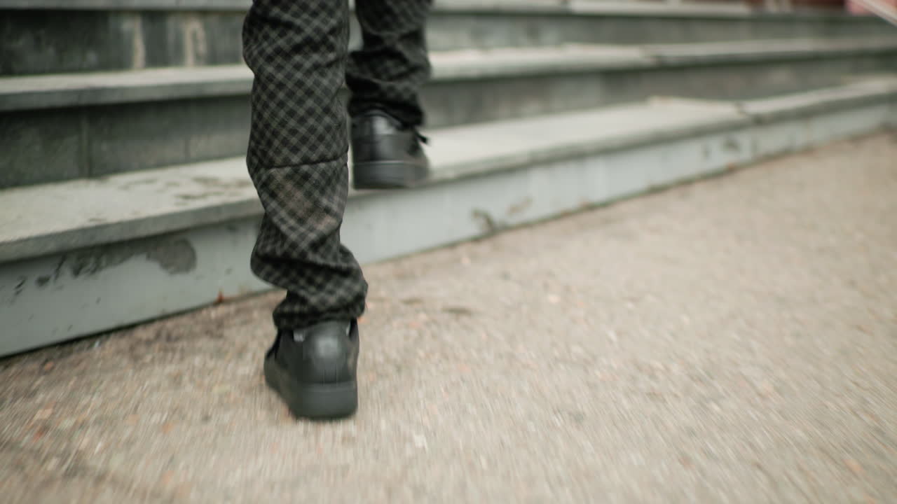 Vista de piernas de una persona con pantalones oscuros y zapatillas deportivas caminando hacia un edificio escolar y subiendo una escalera; flores difuminadas y scooters aparcados visibles al fondo.