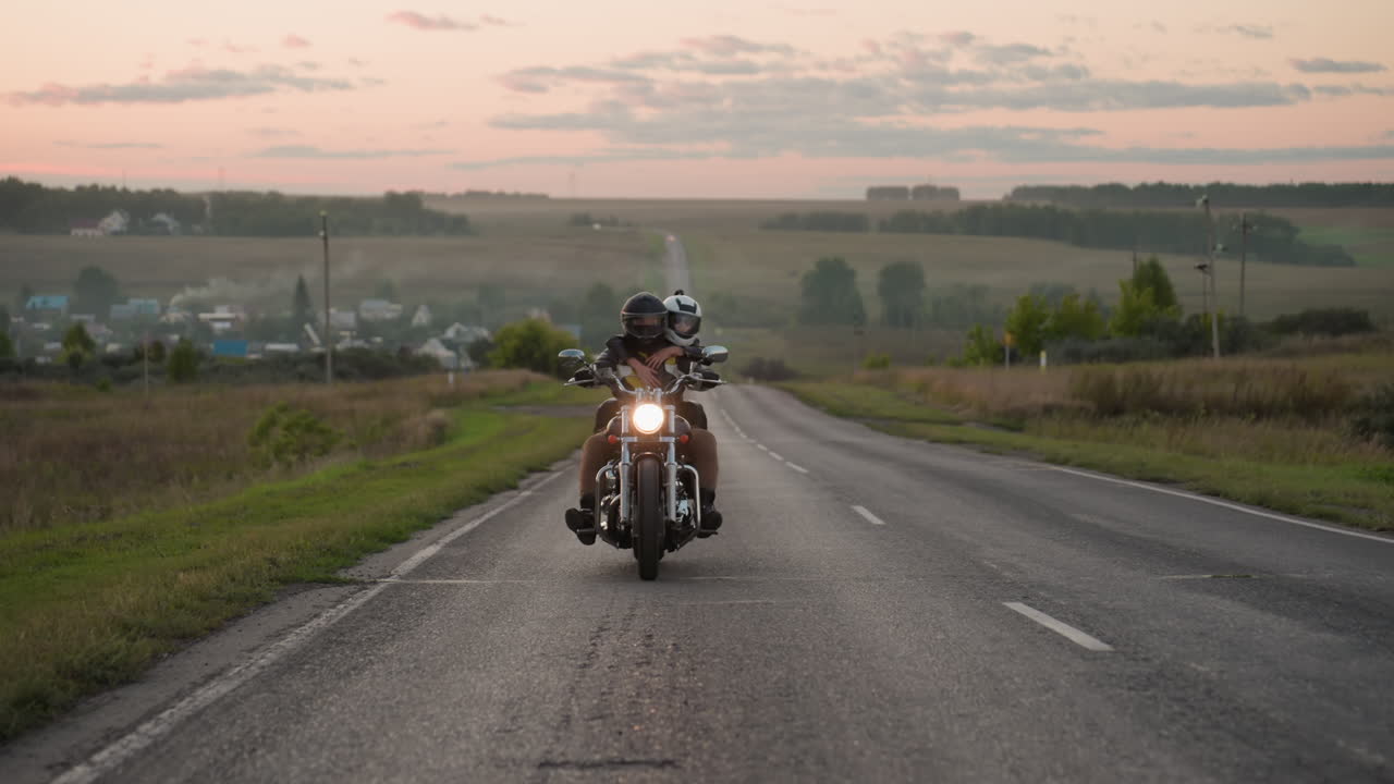 Motorcycle rider with passenger wearing helmets drives along empty countryside road during sunset, glowing headlight bright against scenic rural landscape with fields, trees, and soft evening horizon