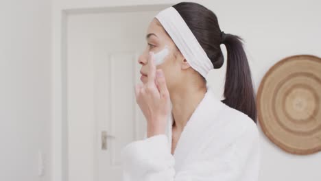 biracial woman applying cream on face in bathroom