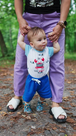 Little toddler stepping slowly looking around. Mommy's holding her baby helping him to walk. Nature blurred backdrop. Vertical video