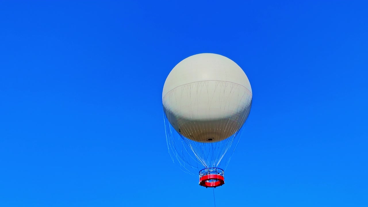 Balon udara panas mengapung di langit biru cerah pada hari yang cerah di atas taman, menawarkan pemandangan indah dan petualangan unik bagi pengunjung di bawah.