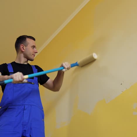 Painter man at work painting a wall with a roller. Worker in blue overalls doing renovation of a room. Offering professional painting services. View from below