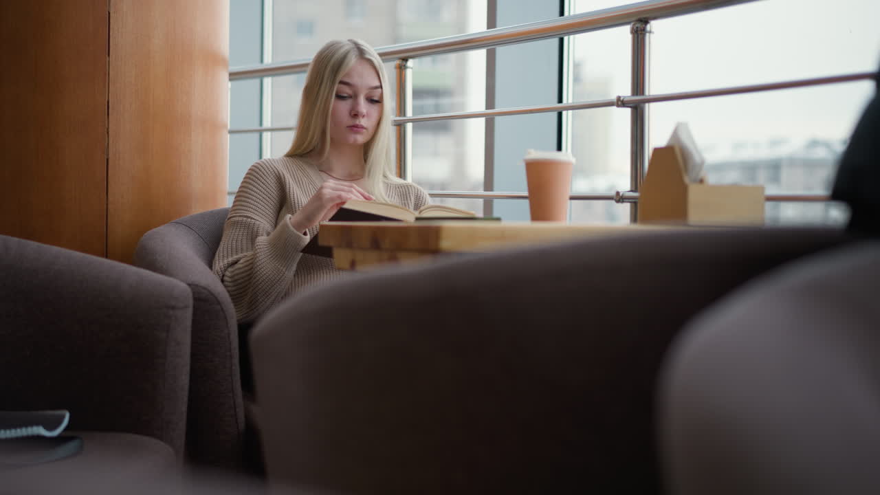 테이블에 앉아있는 여인이 책 읽고, 편안하고 현대적인 카페 환경에서 테이블 위에 커피 컵과 킨을 가지고 집중하는 동안 단 하나의 페이지를 부드럽게 들어 올립니다.