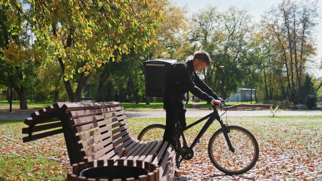 Delivery cyclist autumn park approaches bicycle, mounts bike with insulated backpack, pedals along leaf covered path beside wooden bench, sunlight filtering through trees, urban food courier completes