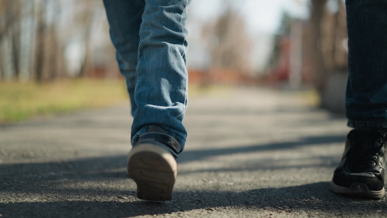 un primer plano de un niño con pantalones vaqueros y botas caminando por una acera con alguien, el niño está levantando una pierna en medio del paso