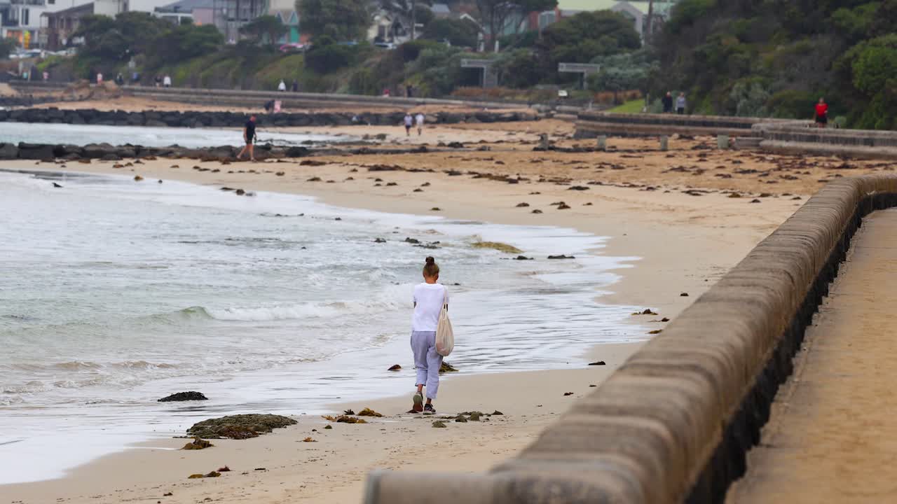 A person walks along a sandy beach beside a seawall under overcast skies at Point Lonsdale, Australia