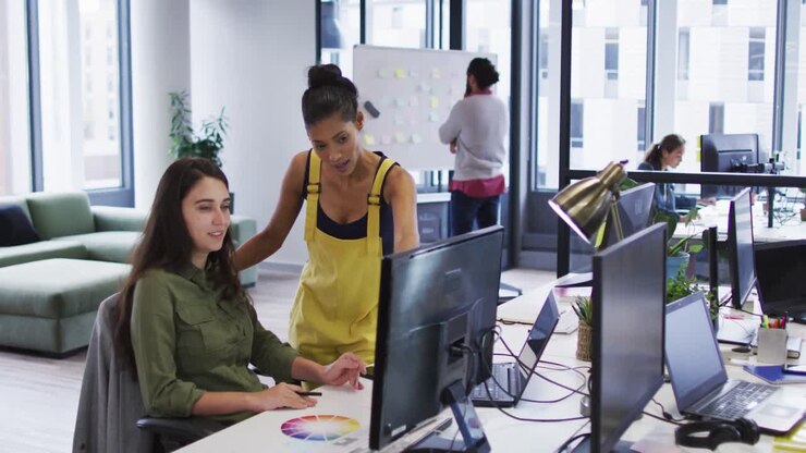Two diverse female work colleagues looking at computer monitor and talking
