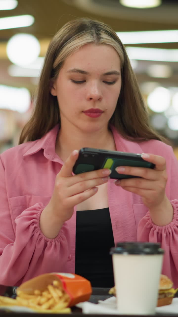 dama blanca en vestido rosa sentada en una mesa de madera en un restaurante ocupado, ajustándose para tomar una foto de su comida, la comida consiste en una hamburguesa, papas fritas y una taza de café en una bandeja negra