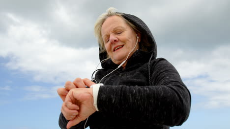 Low-angle-view-of-old-caucasian-senior-woman-using-smartwatch-and-listening-music-on-earphones-at-be