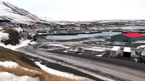 Húsavík-Town-Village-in-Norðurþing-Municipality,-North-Coast-of-Iceland-in-Winter,-View-of-the-Local-Port,-Houses-on-Edge-of-Snowy-Mountain,-Warehouse-and-Empty-Road,-Coastal-Landscape-View