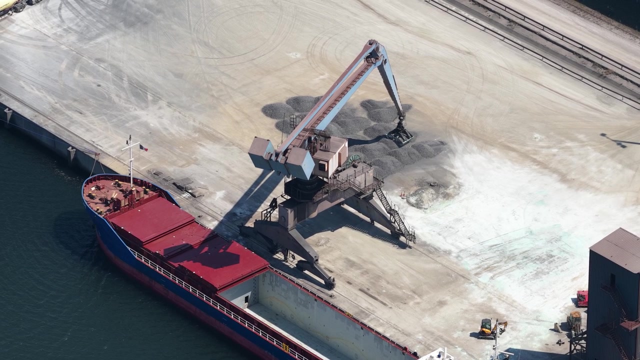 Industrial crane at a seaport loading raw materials like gravel or stone from the quay into the open hold of a large bulk carrier ship docked in the harbor, filmed from an aerial perspective