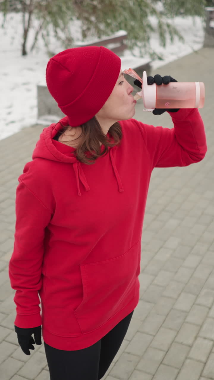lady in red hoodie and beanie drinking from pink water bottle while standing outdoors in snow-dusted park, surrounded by trees, serene winter atmosphere, and red bag placed on nearby bench