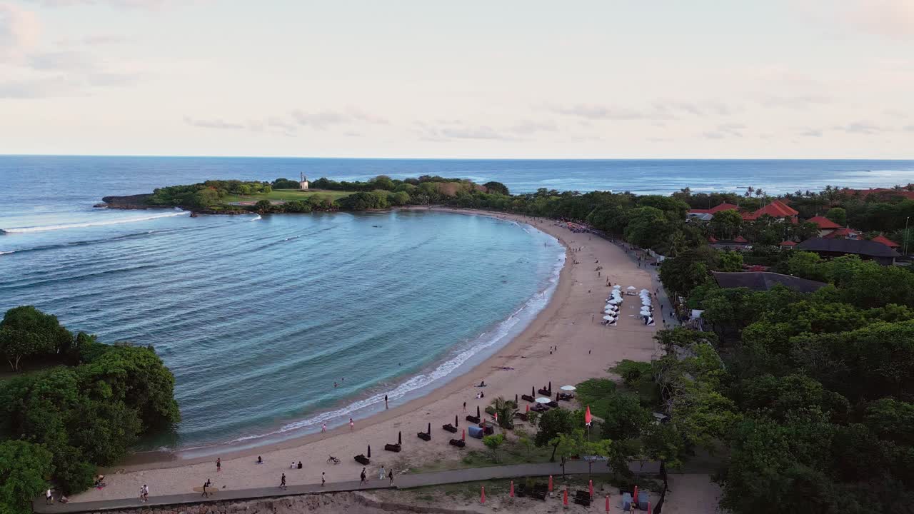 Drone video showing a tropical coastline with white sand beach, calm shallow water, beach umbrellas, gentle evening light and a resort area stretching along the curved shoreline in Nusa Dua Bali