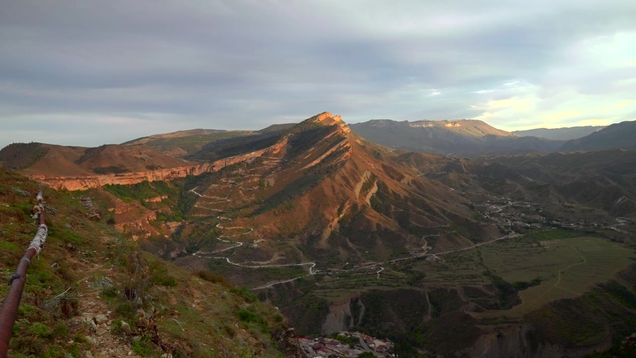 Panorama de montanha elevado exibe um cume nítido e ensolarado, vale profundo, estradas sinuosas, casas de vilarejo dispersas e campos verdes abaixo, com riqueza de detalhes de montanha, cume, vale e paisagem.