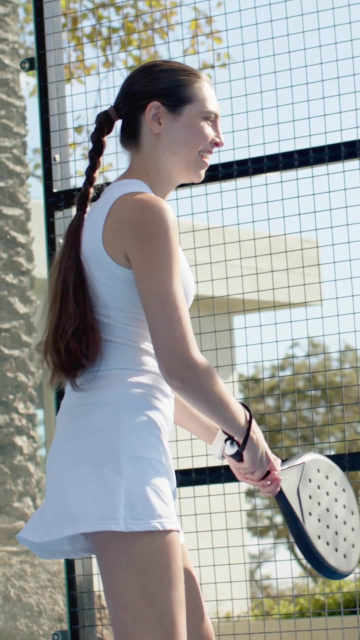 Vertical video: Smiling woman in white dress, adjusting paddle while opponent hitting shot on court