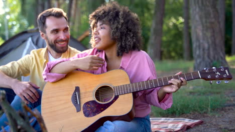 happy hiker couple camping in the forest. young black woman plays guitar and sings, caucasian man smiles.