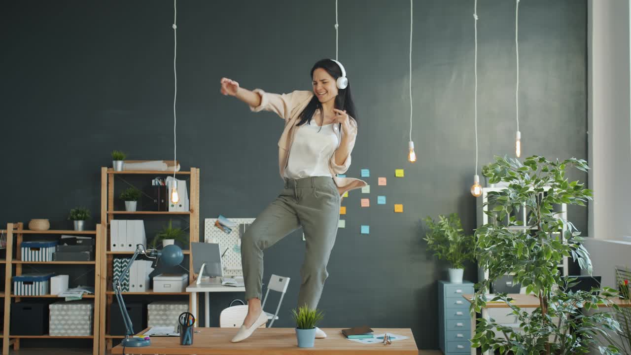 Affarista felice che balla sulla scrivania dell'ufficio, scherzando e godendosi la musica in cuffia