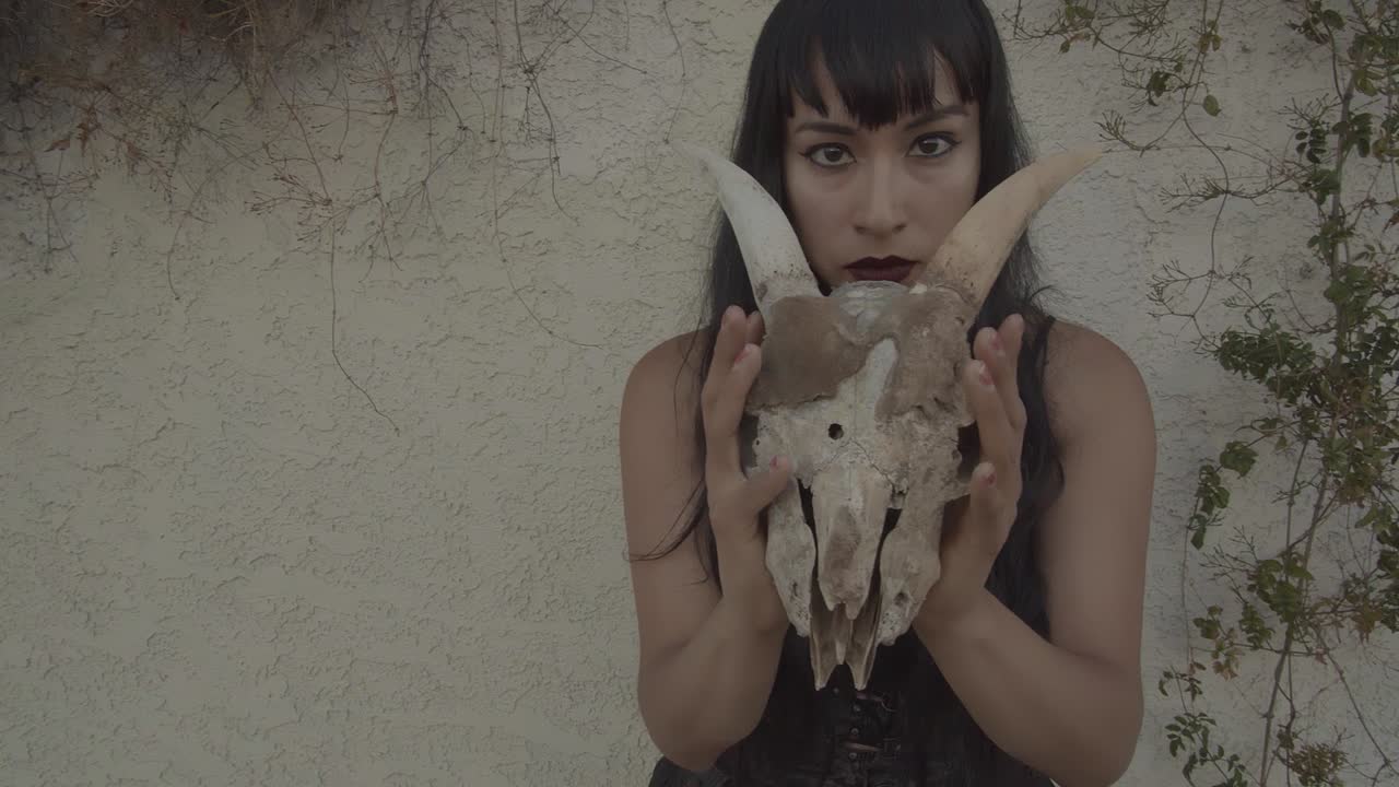 Zoom out of girl with long black hair and bangs, rotating an animal skull with horns in her hands
