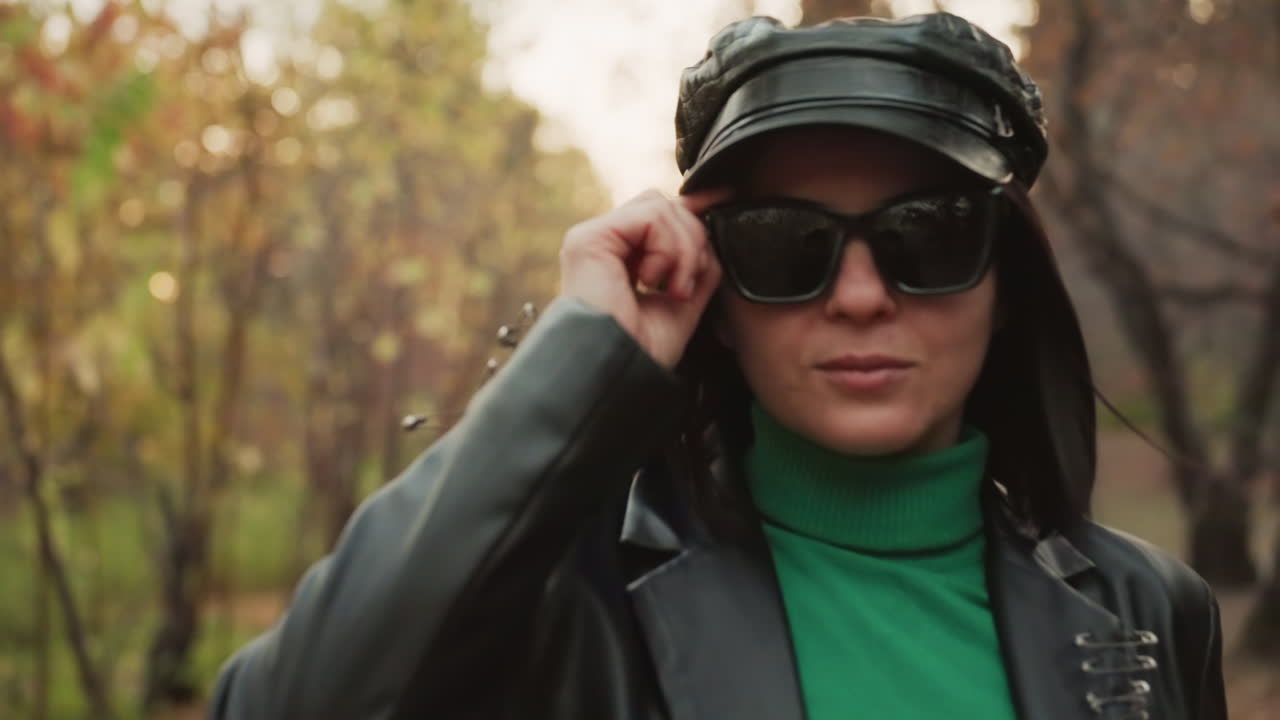 woman in leather jacket approaching camera on treelined path, stylish reveal as she lifts sunglasses, editorial fashion blogger pose with glossy cap, warm autumn light and subtle bokeh, bold attitude