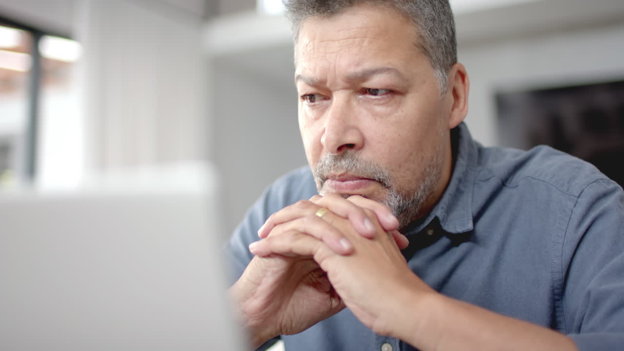un homme biracial sénior concentré assis à table en train de regarder un ordinateur portable, travaillant à domicile, au ralenti.