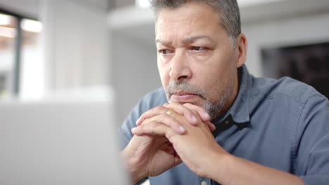 hombre biracial sénior centrado sentado en la mesa mirando la computadora portátil, trabajando desde casa, en cámara lenta
