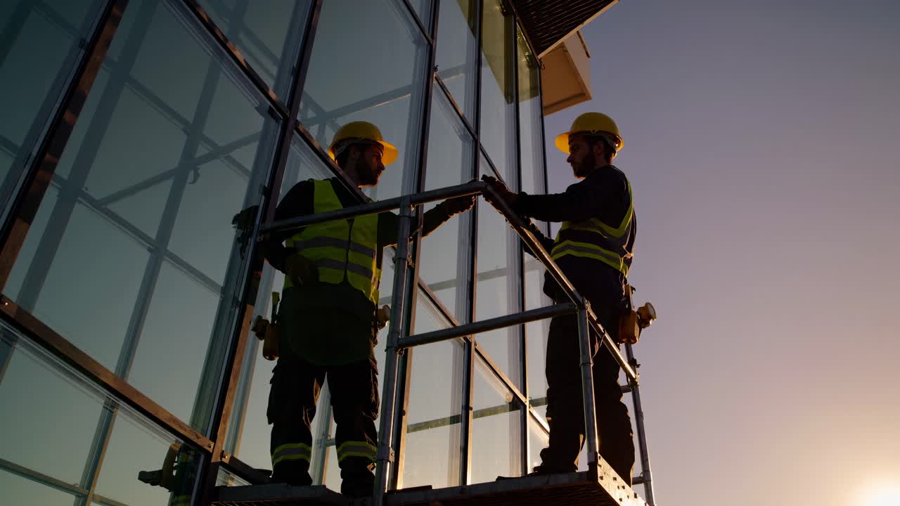 Silhouetted workers in safety gear on scaffolding at sunset, captured from a low angle