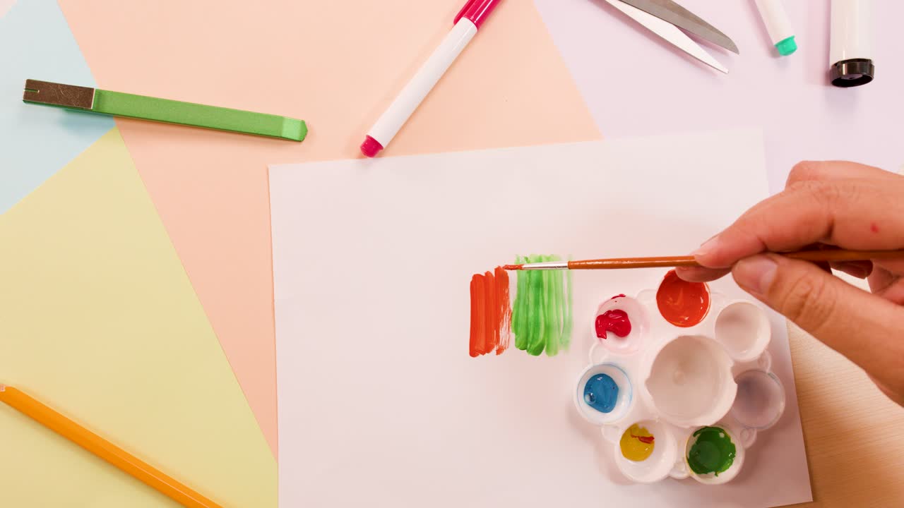 Hand uses paintbrush to apply orange, green, yellow stripes on white paper, overhead view