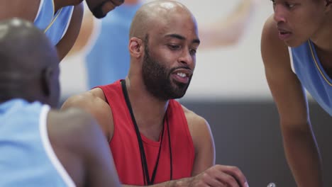 diverse male basketball team and coach with clipboard in discussion at indoor court, in slow motion