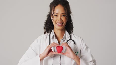 Video-of-portrait-of-smiling-biracial-female-doctor-holding-heart-on-white-background