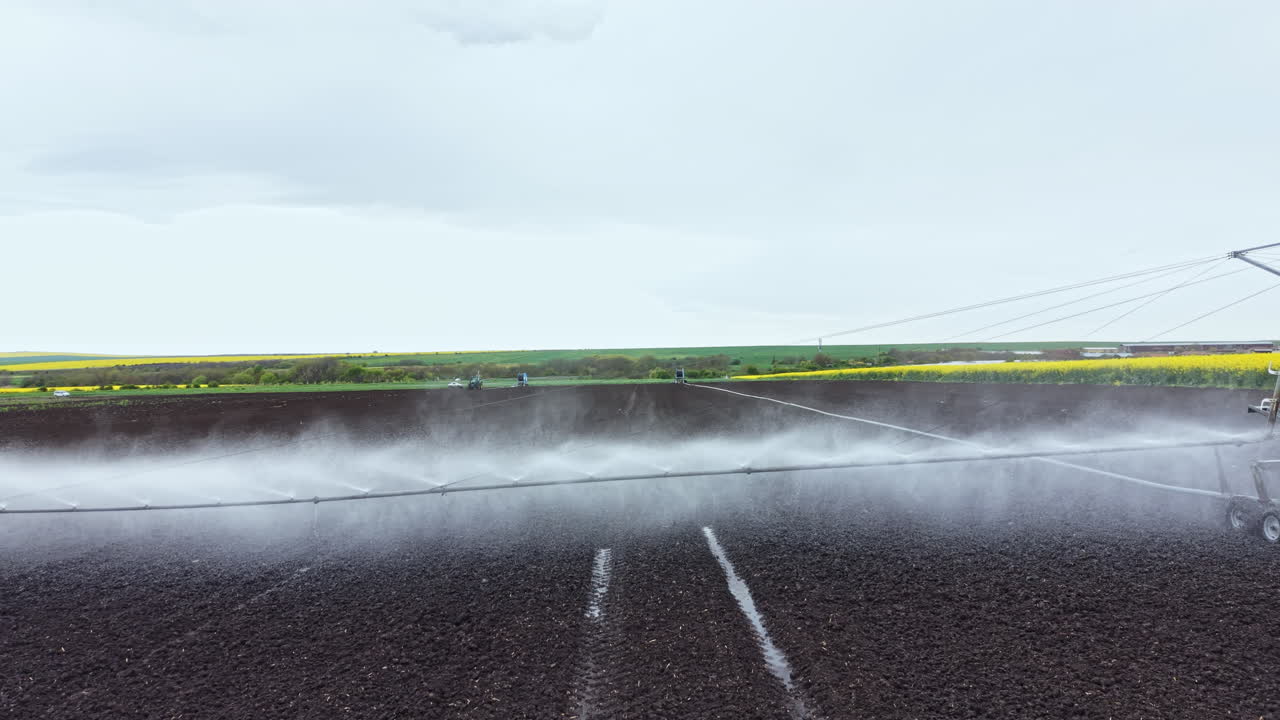 Sistema di irrigazione che annaffia un campo durante una giornata nuvolosa in un'area agricola con paesaggio verde sullo sfondo.