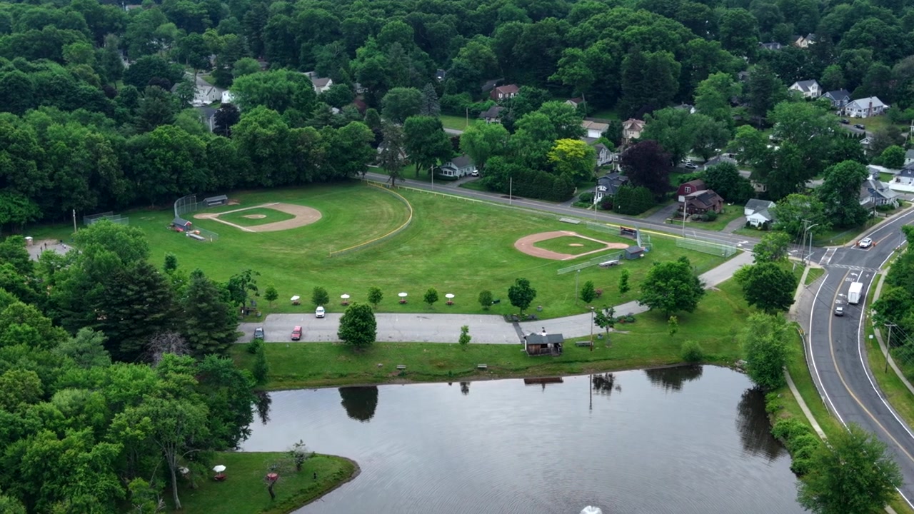 Drone mengorbit tepat di atas taman East Longmeadow yang menampilkan dua lapangan bisbol, kolam yang indah, jalan-jalan perumahan, dan pepohonan hijau rimbun di Massachusetts, AS