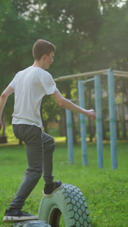 a young boy in a white shirt performs outdoor training in slow motion, jumping across tires on a grassy field, the background features trees and various sports equipment