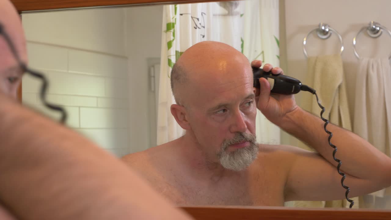 Caucasian male using electrical trimmer to cut his beard in a bathroom. Back view from behind, reflection in mirror, close up