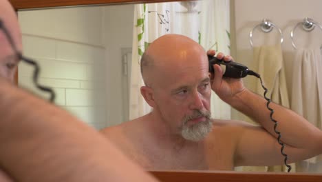 caucasian male using electrical trimmer to cut his beard in a bathroom. back view from behind, reflection in mirror, close up