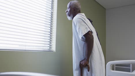Afroamerikanischer-Männlicher-Patient,-Der-Im-Krankenhauszimmer-Mit-Einem-Stock-Zum-Fenster-Geht