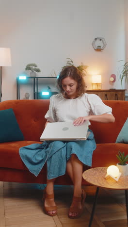 happy young woman lady sitting on sofa opens laptop and working on project in living room at home