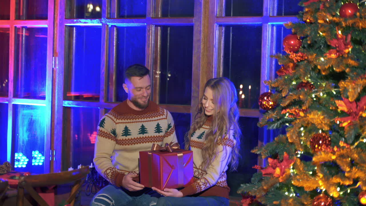Couple in love near Christmas tree. Handsome man giving his girlfriend a big gift in red box for Christmas. Romantic atmosphere at home. New Year time.