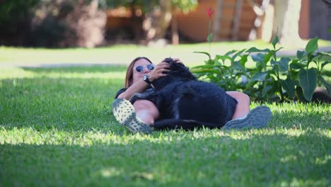 a young caucasian woman lying on the grass plays with her black dog in the garden of a house on a sunny day