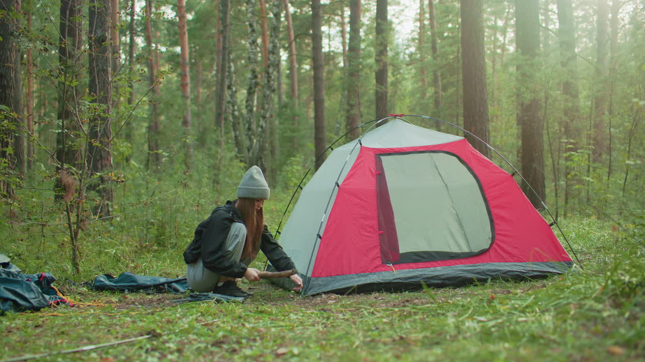 camper with long hair pegs tent by pressing stake into ground using large stick while crouching beside pitched red and gray shelter in forested campsite surrounded by tall trees and green vegetation