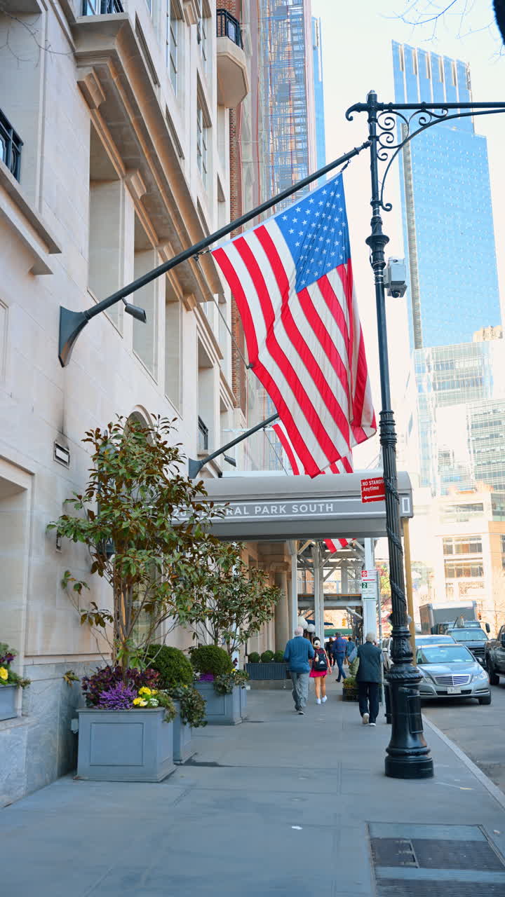 New York, USA, 14 marzo 2026: Bandiera americana sulla facciata di un edificio lungo Central Park South. Primo piano della bandiera USA appesa sopra un marciapiede con pedoni e un classico lampione di New York