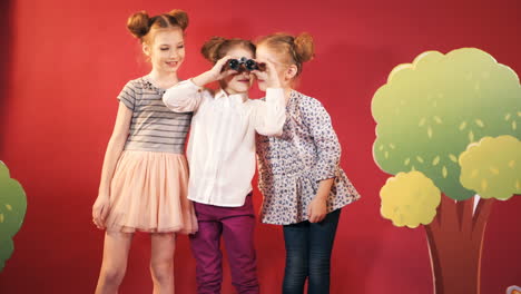 three happy girls playing together with binoculars on the red background. pretty little girls looking into the black binoculars in childish studio with trees decor.